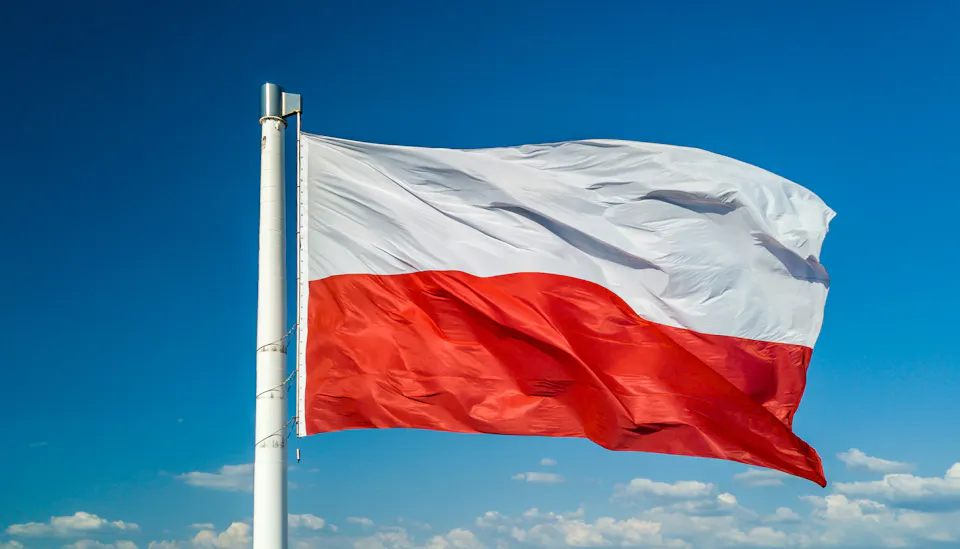 Red and white Polish national flag waving on a white metal pole against a clear blue sky with scattered clouds.