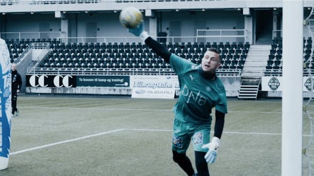 Goalkeeper in green kit reaching up to catch a ball at an outdoor stadium with advertising boards and empty stands behind him