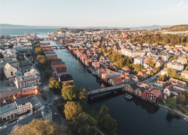 Widok z lotu ptaka na norweskie miasto z szeroką rzeką, kolorowymi budynkami nad wodą, mostami i zielonymi wzgórzami w tle.