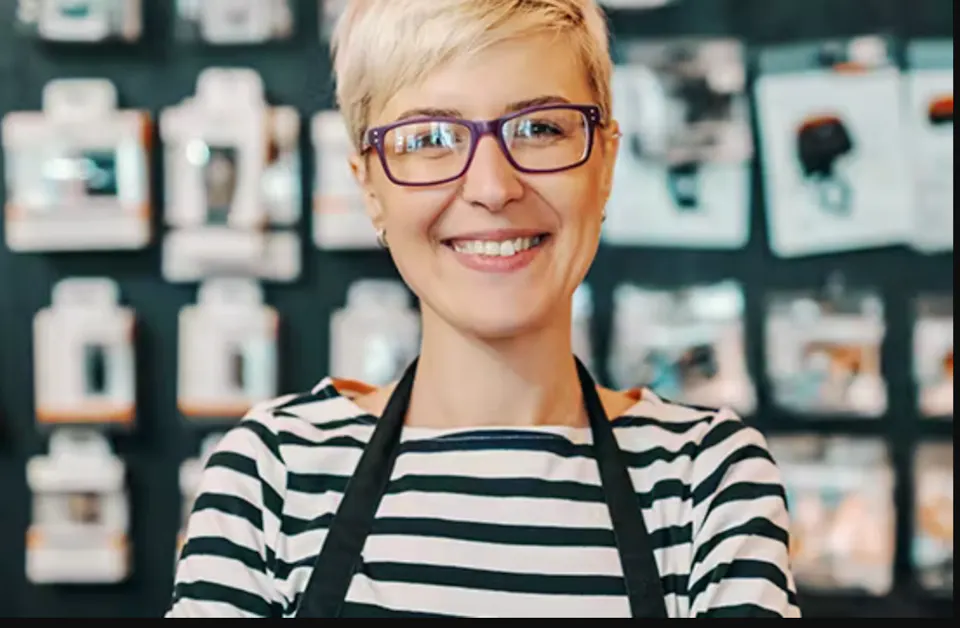 A smiling shop assistant with short blonde hair, purple glasses and a black apron stands in front of a blurred product display wall.