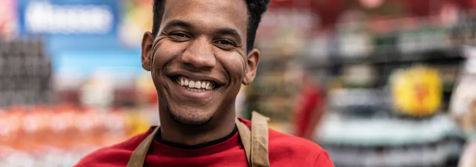 Close-up of a smiling young man in a red shirt with a strap over his shoulder, against a blurred colourful background