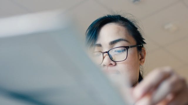 Bliskie zdjęcie portretowe kobiety w okularach i z ciemnoniebieskimi refleksami we włosach, patrzącej w dół na urządzenie, z rozmytym pierwszym planem.