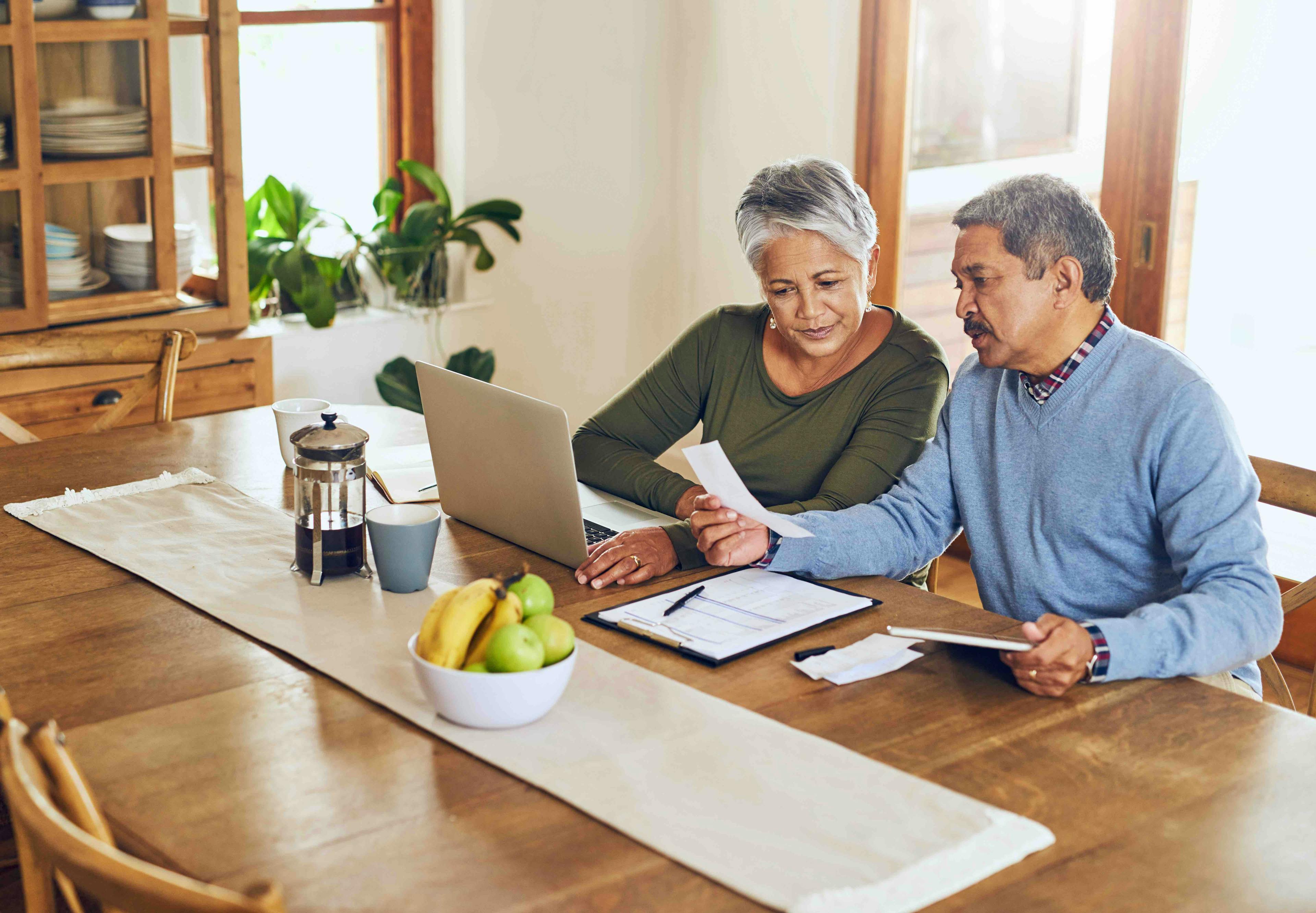 An older Hispanic husband and wife sit at the table in front of a laptop and do their taxes
