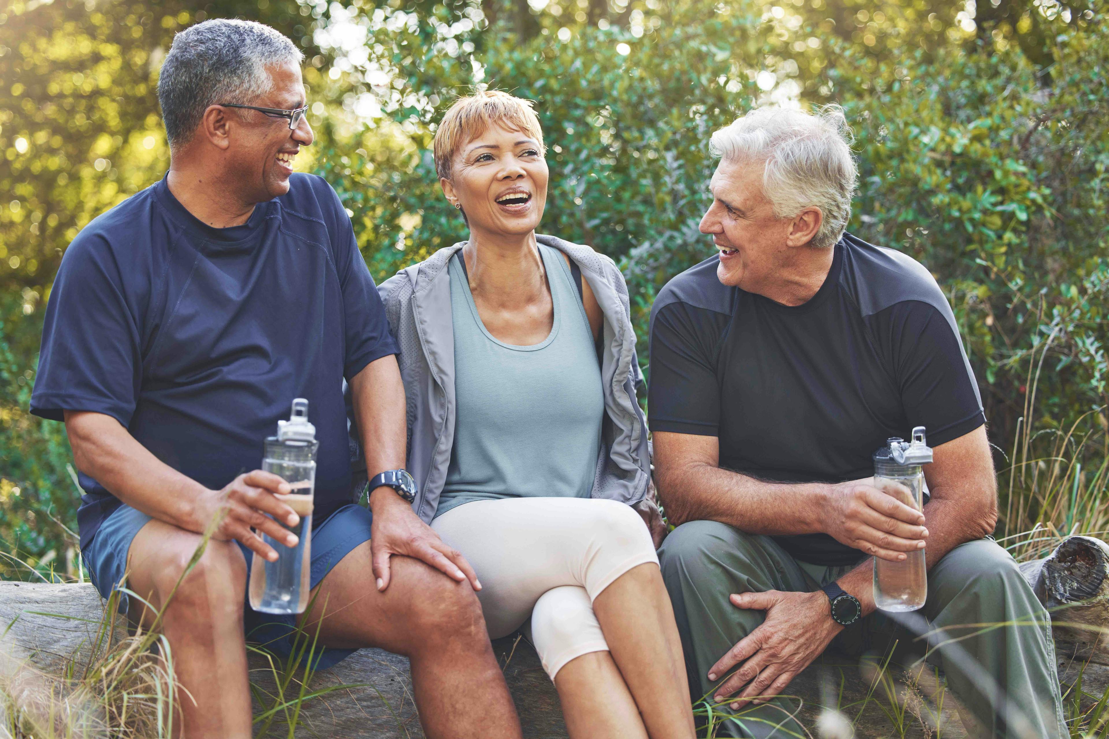senior friends in conversation and drinking water while sitting in the forest after hiking