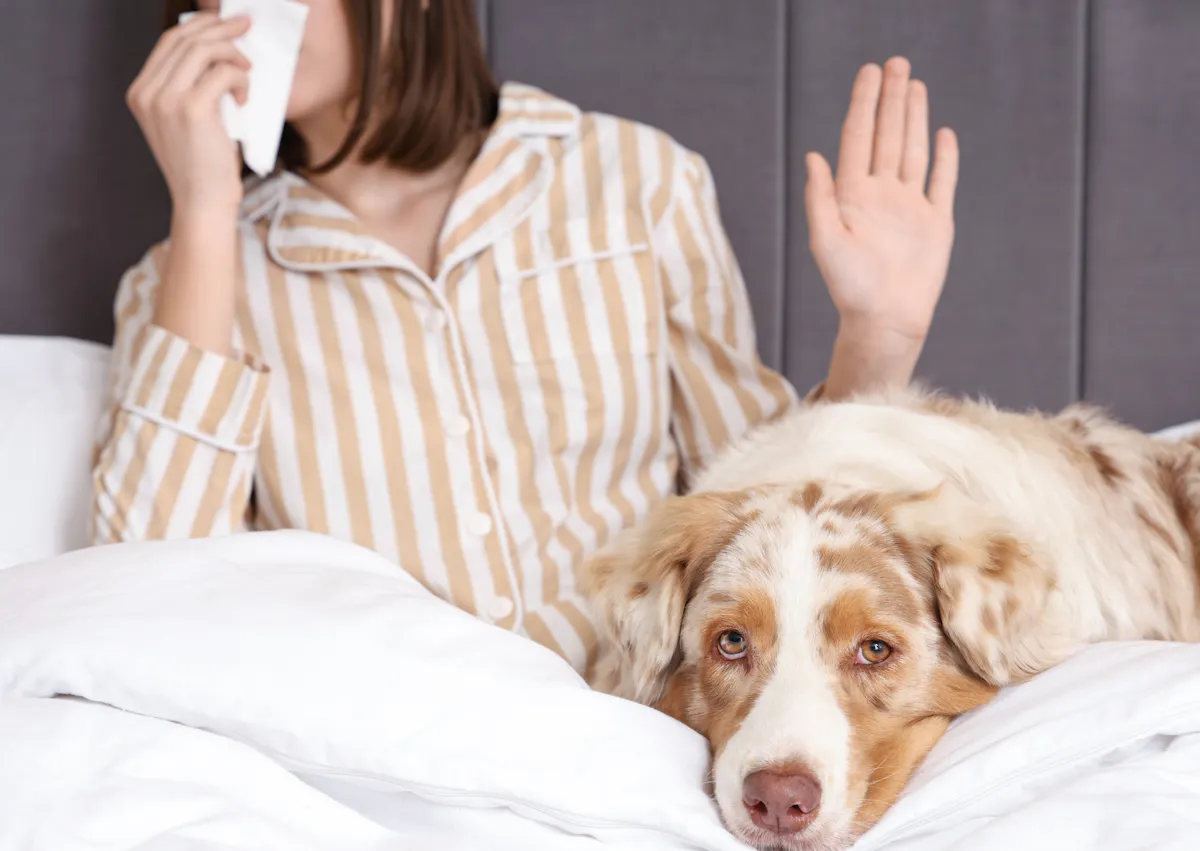 une femme fait stop de la main et se mouche, pendant que son chien est couché dans le lit 