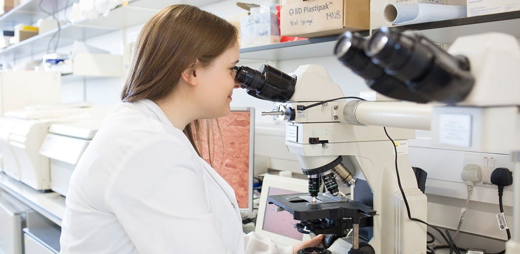 Scientist in a lab with a lab coat, looking into a microscope.
