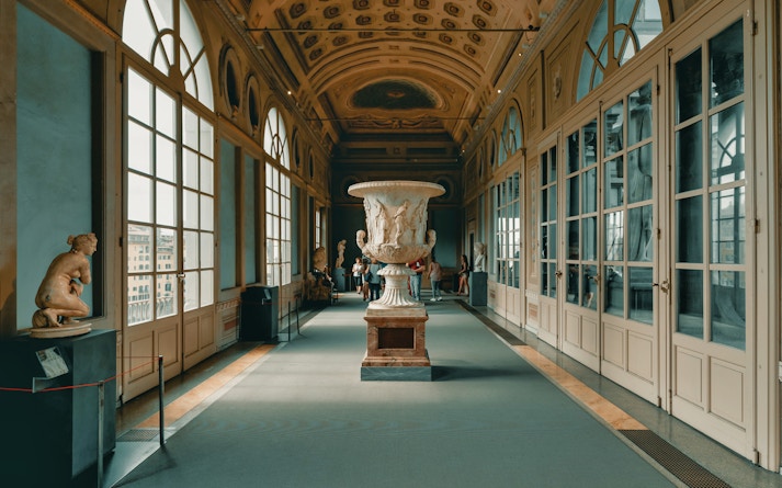 Spacious corridor inside Uffizi Gallery with large windows overlooking Florence, featuring a classical sculpture in the center and visitors enjoying the artworks