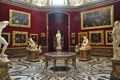 Intimate second-floor room in the Uffizi Gallery, with marble sculptures set against luxurious red walls and golden-framed artworks