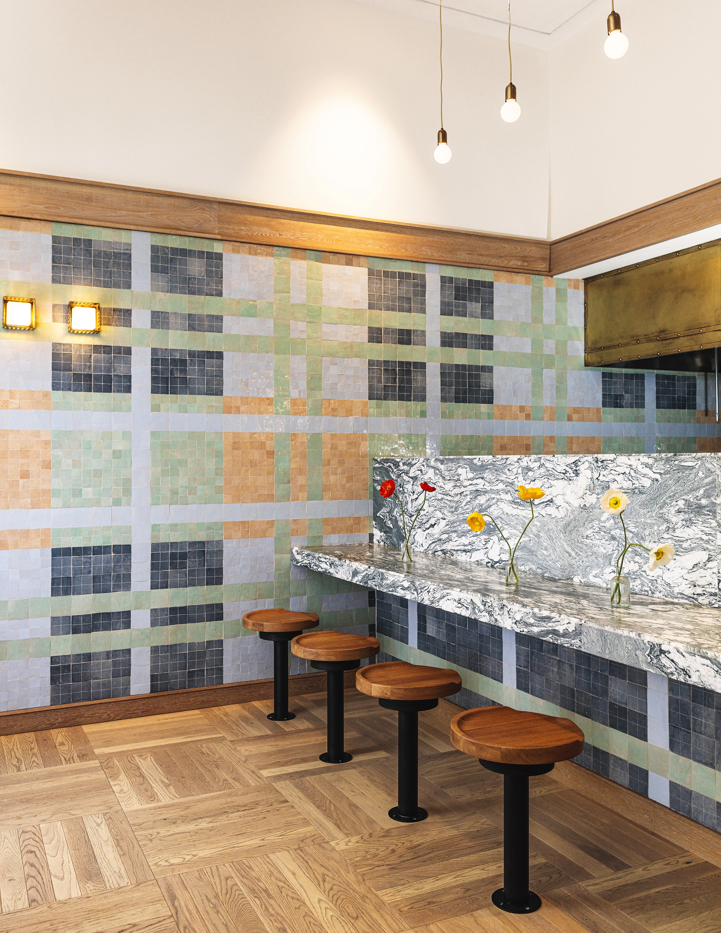 Stylish restaurant interior at Austin Proper Hotel and Residence, featuring a plaid-patterned tiled wall, a sleek marble countertop, and wooden stools with black metal bases. The space is illuminated by modern pendant lighting, while fresh flowers add a touch of elegance to the design.