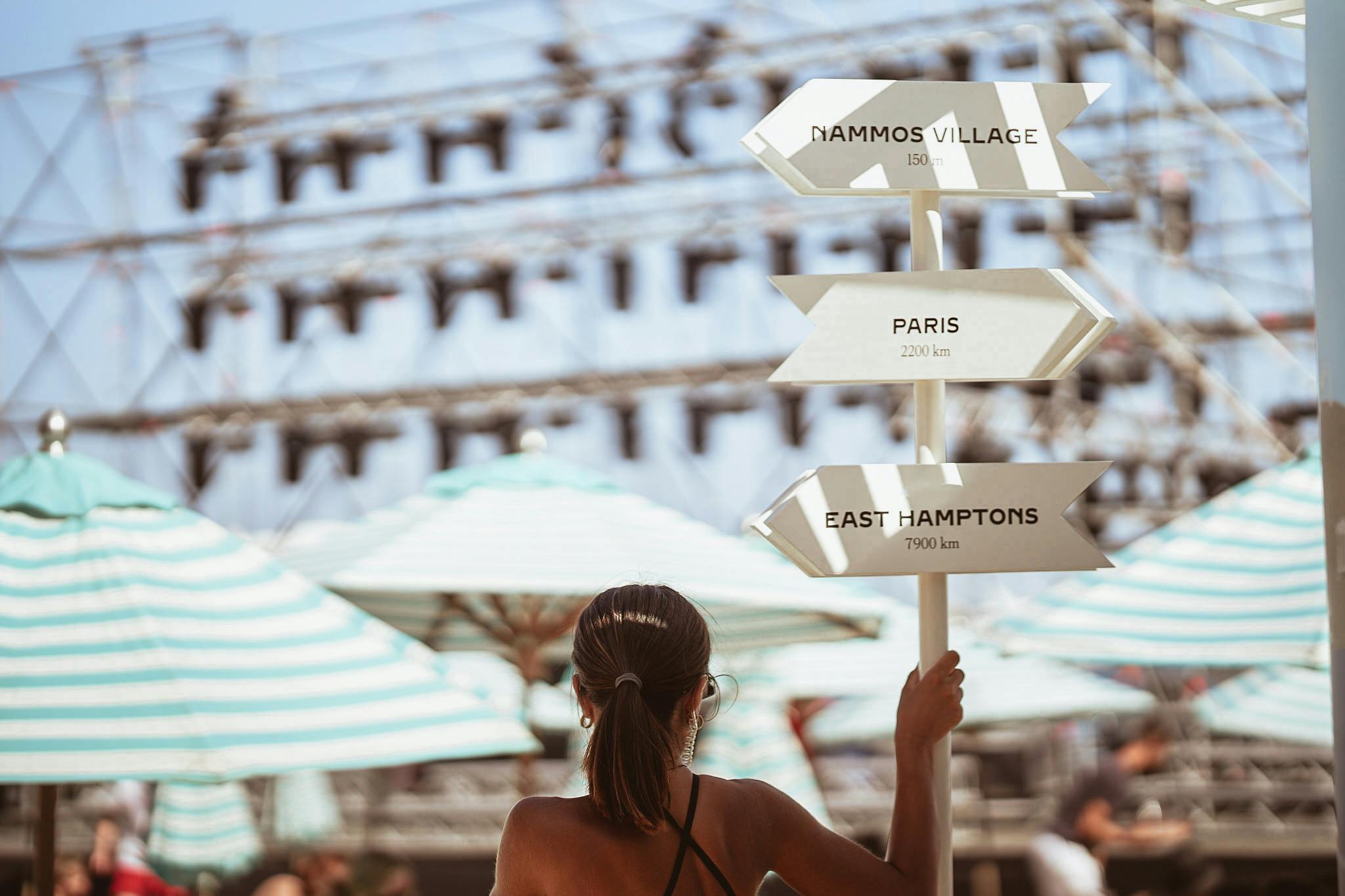 Lady next to directional sign showing amount of kilometres to Nammos Village, Paris and East Hamptons