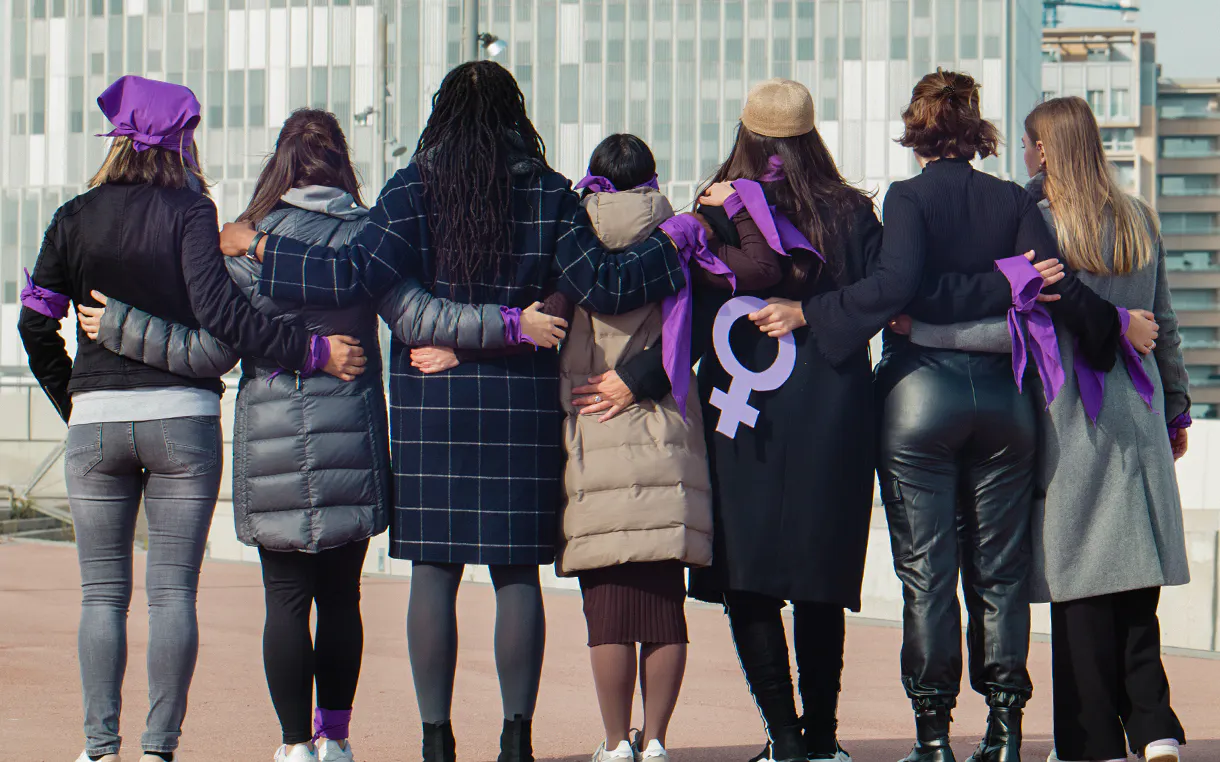 A group of diverse woman holding each other facing away and looking out at a city