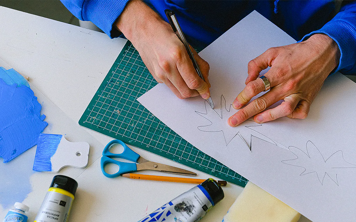 Hands cutting out a star shape from paper using an X-Acto knife. Art supplies are spread across the table.