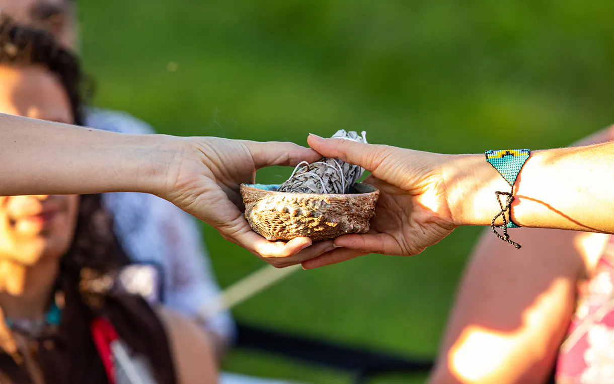 Two hands pass along an abalone shell with a sage bundle in it.