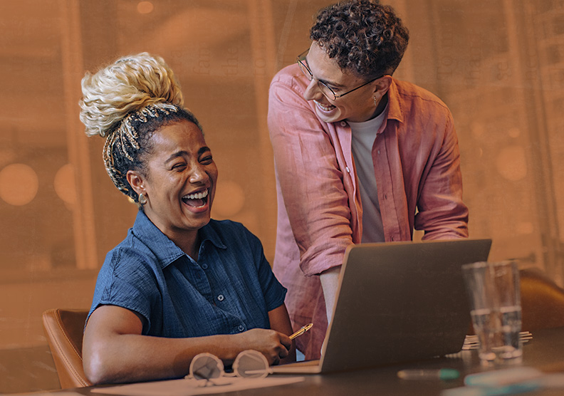 A person in a pink button up is standing near their colleague that is working on their laptop and desk. These two professionals are laughing together in their modern workplace. 