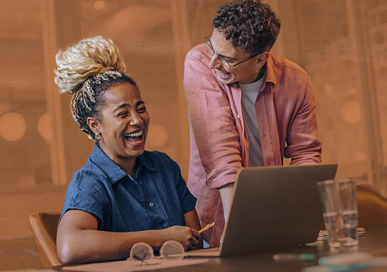 A person in a pink button up is standing near their colleague that is working on their laptop and desk. These two professionals are laughing together in their modern workplace.