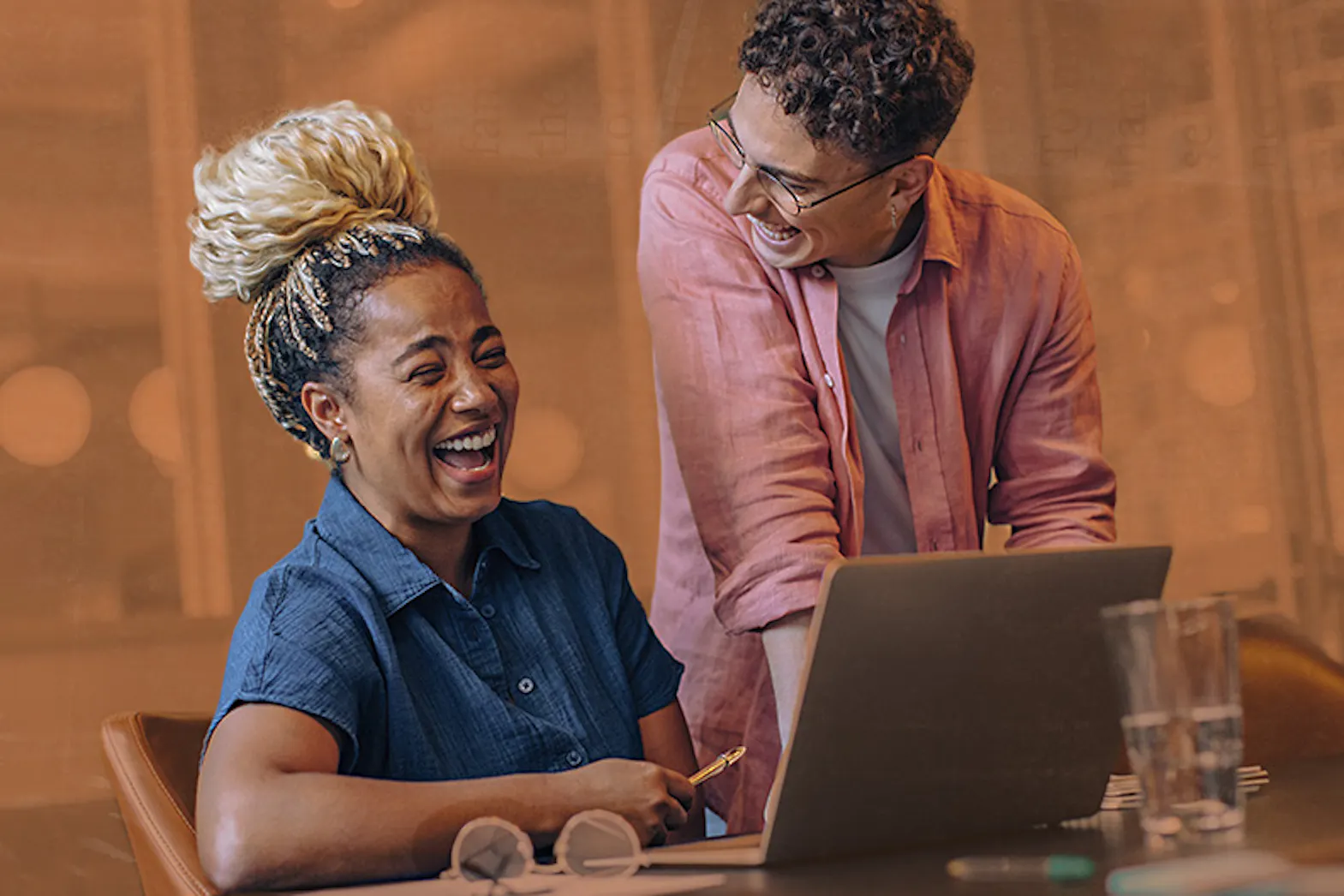 A person in a pink button up is standing near their colleague that is working on their laptop and desk. These two professionals are laughing together in their modern workplace.