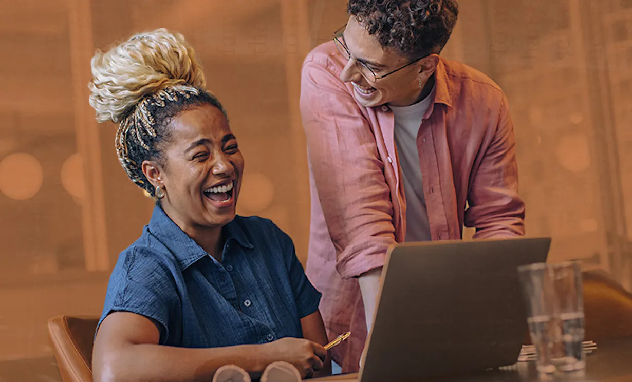 A person in a pink button up is standing near their colleague that is working on their laptop and desk. These two professionals are laughing together in their modern workplace.