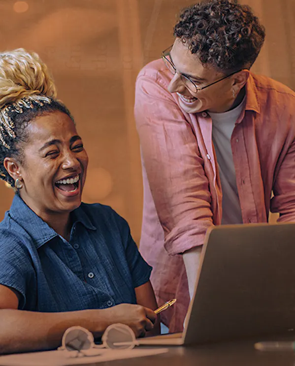 A person in a pink button up is standing near their colleague that is working on their laptop and desk. These two professionals are laughing together in their modern workplace.