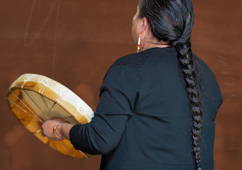 A woman with long, braided hair holds a drum while singing.