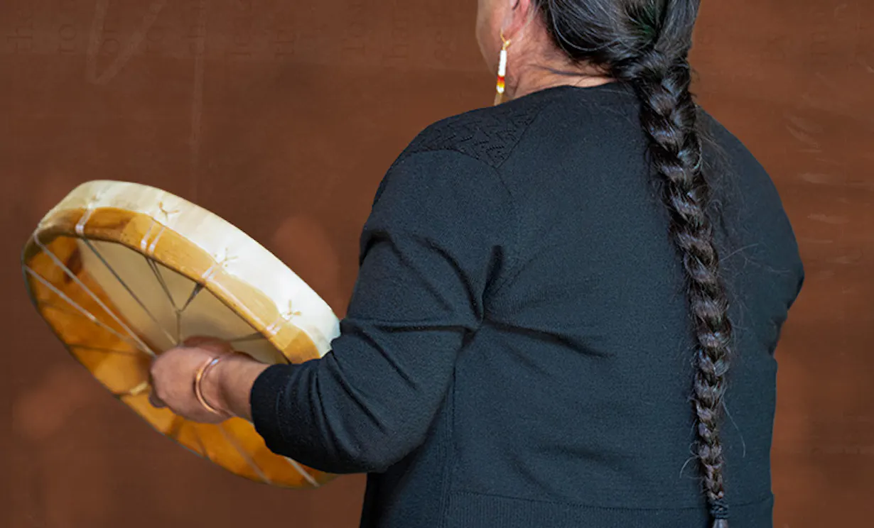 A woman with long, braided hair holds a drum while singing.