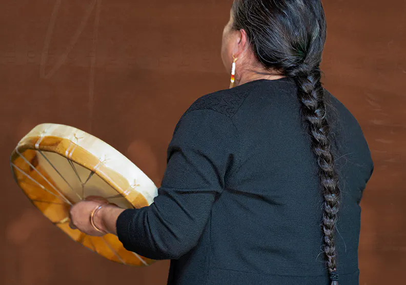 A woman with long, braided hair holds a drum while singing.