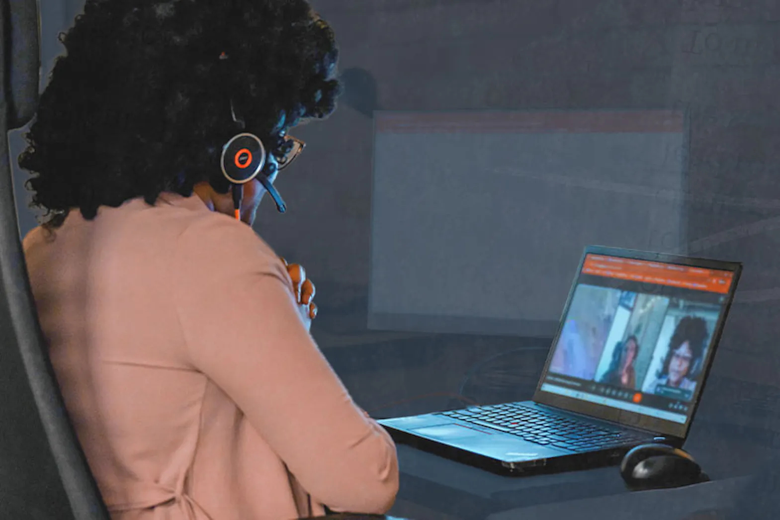 A woman sits at a desk looking at a computer screen. The screen shows a remote meeting.