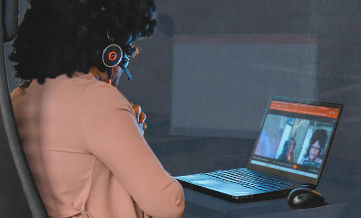 A woman sits at a desk looking at a computer screen. The screen shows a remote meeting.