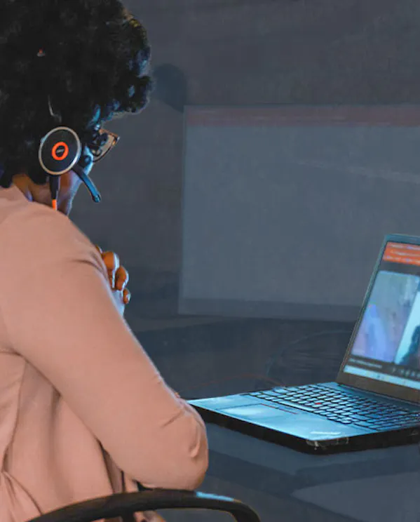 A woman sits at a desk looking at a computer screen. The screen shows a remote meeting.
