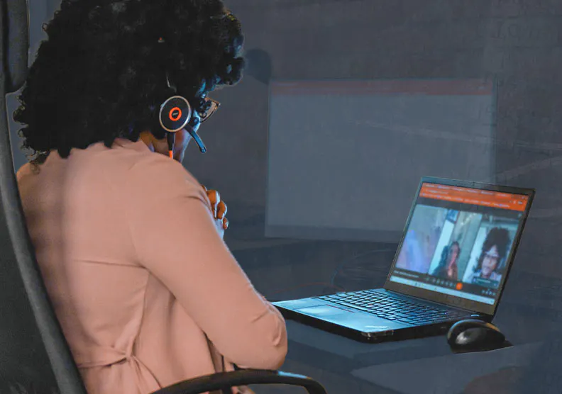 A woman sits at a desk looking at a computer screen. The screen shows a remote meeting.