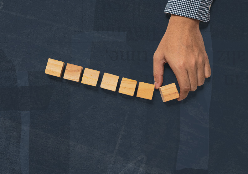 An overhead view shows a person wearing a blue checkered shirt placing a small wooden block at the end of a row. Seven wooden cubes sit neatly aligned across a textured dark blue surface.