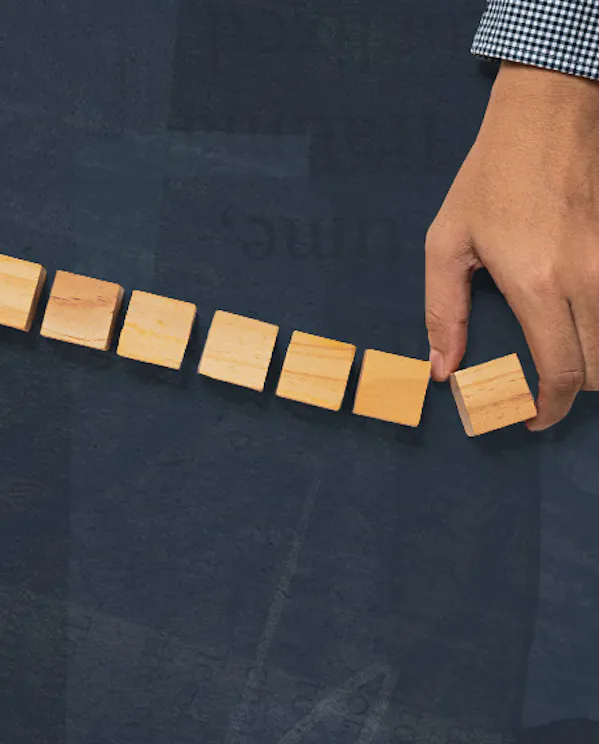 An overhead view shows a person wearing a blue checkered shirt placing a small wooden block at the end of a row. Seven wooden cubes sit neatly aligned across a textured dark blue surface.