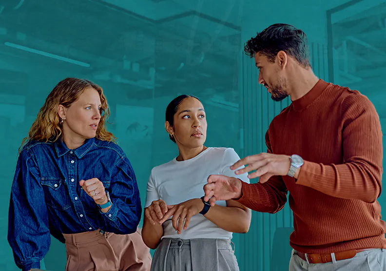 A diverse group of three professionals actively communicating in a modern workplace setting. A man in a brown sweater gestures to convey a point while two female colleagues listen attentively, modeling workplace conflict transformation, operational trust, and flexible governance in team planning.