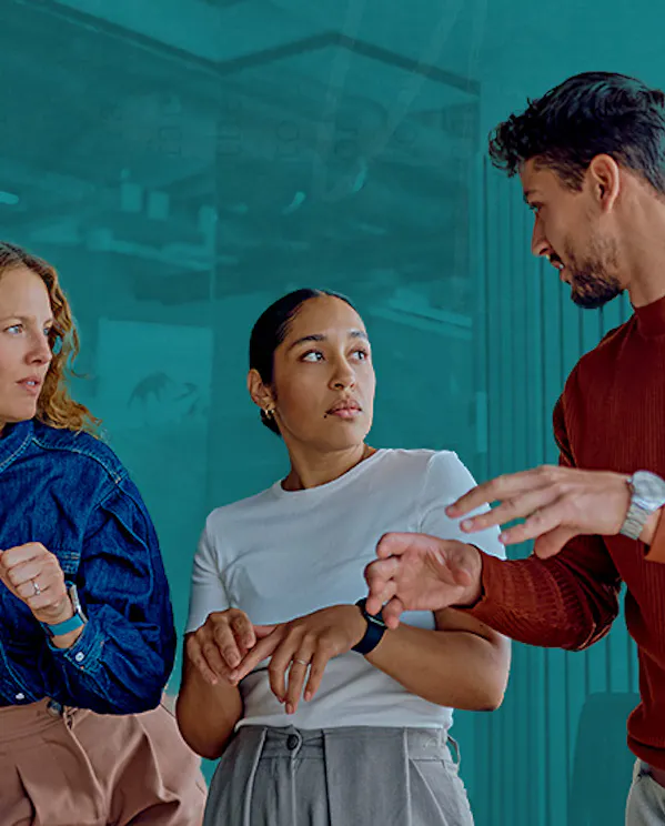A diverse group of three professionals actively communicating in a modern workplace setting. A man in a brown sweater gestures to convey a point while two female colleagues listen attentively, modeling workplace conflict transformation, operational trust, and flexible governance in team planning.