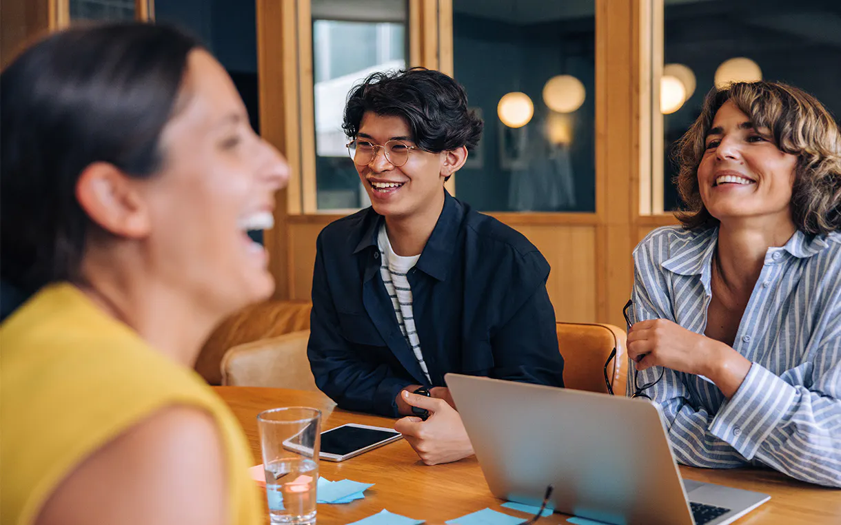 In the foreground, a person in yellow is laughing with two fellow colleagues in a professional workplace setting.