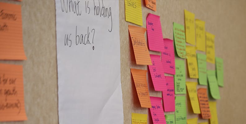 Big white paper that says What is holding us back with multi coloured sticky notes on beige wall