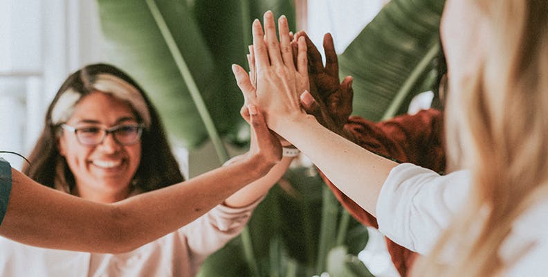 A diverse group of women stand in a circle and high five each other in the center