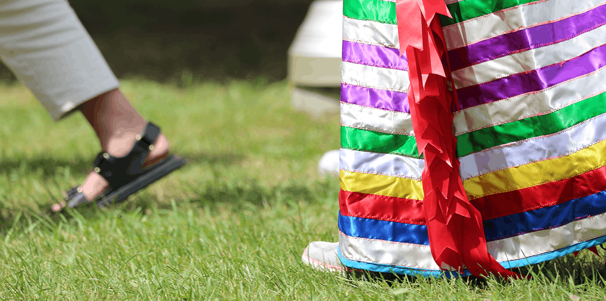 A photo of two peoples feet walking in the grass. One person is wearing black sandals and the other is wearing a long ribbon skit.