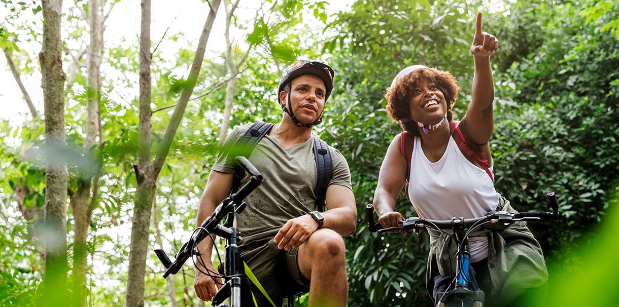 A man and woman cycling together on a forest trail surrounded by trees and greenery.