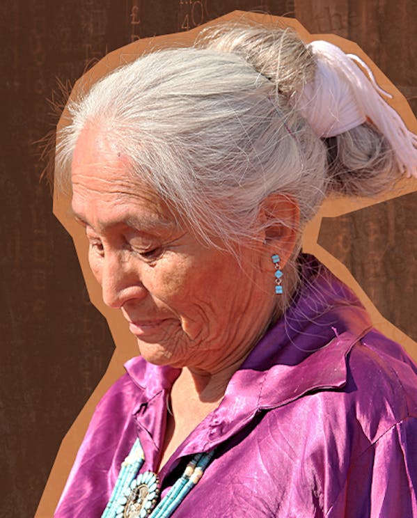 An elder woman dressed in a purple shirt and adorned with a beaded medallion looks down.