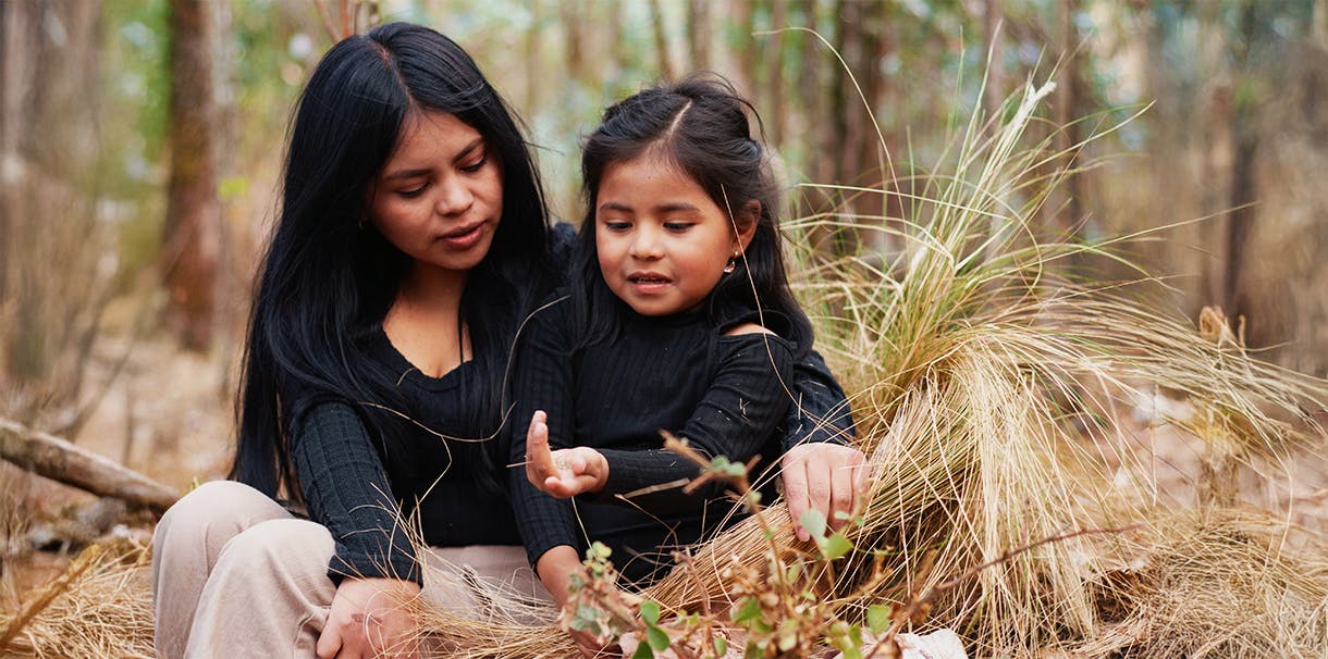 An Indigenous woman and their daughter sit in a forest looking at plants