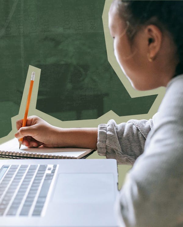 A woman writing on a notebook with a pencil. A laptop is sitting on the desk next to her