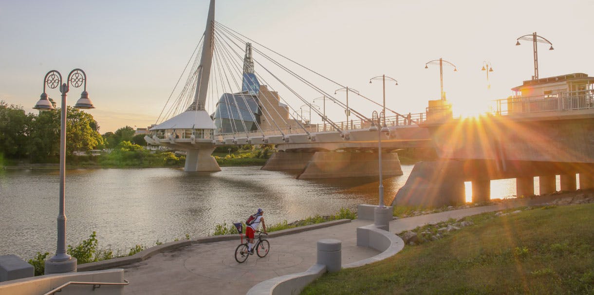 A man riding a bike on a riverside path, showing Winnipeg's skyline at sunset