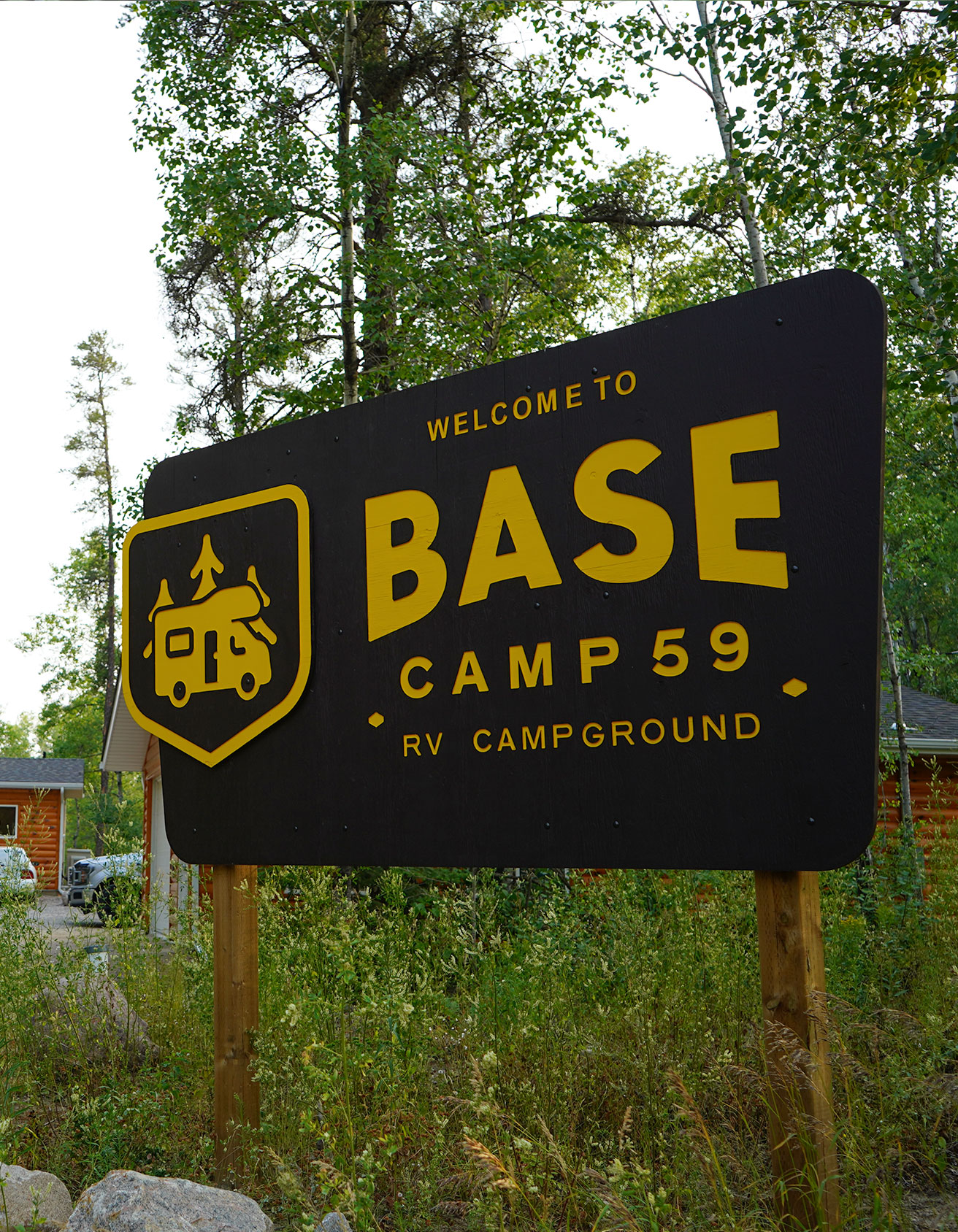 A large, black wooden sign with yellow lettering stands among tall grass and trees, reading "Welcome to BASE CAMP 59 RV CAMPGROUND." To the left of the text, a yellow shield-shaped logo features an icon of an RV with trees on top. In the background, log-cabin-style buildings are partially visible under a clear sky.