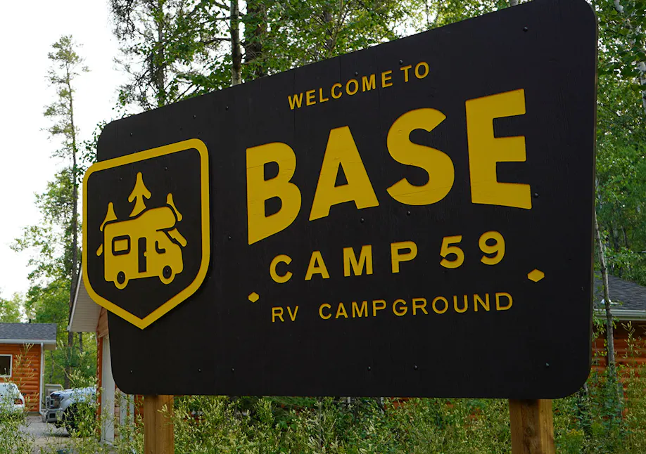 A large, black wooden sign with yellow lettering stands among tall grass and trees, reading "Welcome to BASE CAMP 59 RV CAMPGROUND." To the left of the text, a yellow shield-shaped logo features an icon of an RV with trees on top. In the background, log-cabin-style buildings are partially visible under a clear sky.