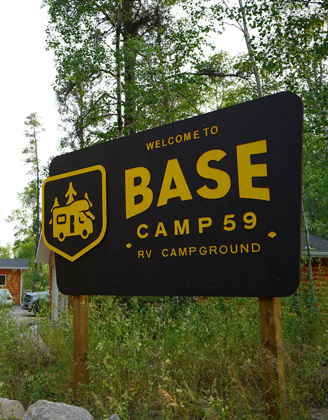 A large, black wooden sign with yellow lettering stands among tall grass and trees, reading "Welcome to BASE CAMP 59 RV CAMPGROUND." To the left of the text, a yellow shield-shaped logo features an icon of an RV with trees on top. In the background, log-cabin-style buildings are partially visible under a clear sky.