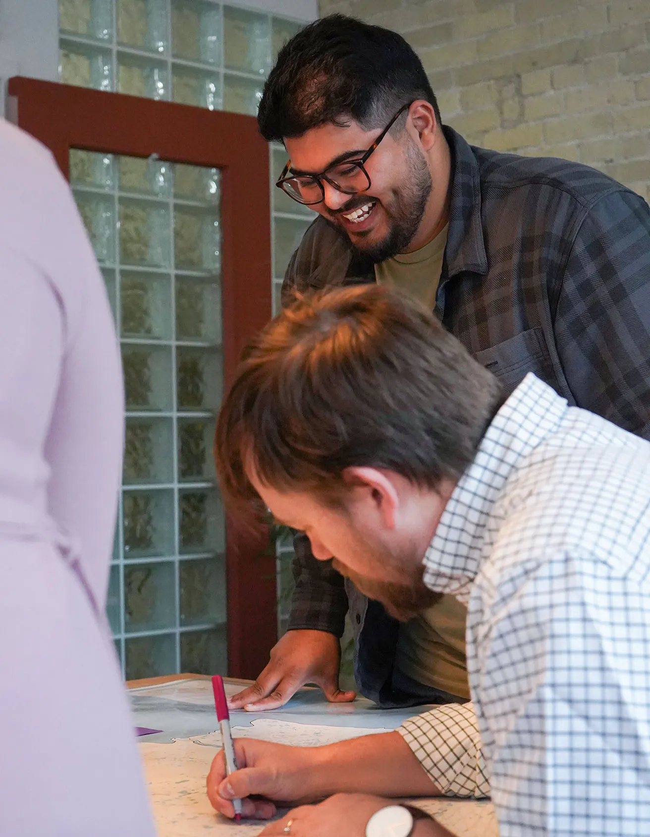 A man with glasses and a beard smiles warmly as he leans over a desk, looking at a large map spread out before him. Beside him, another man in a checkered shirt writes on the map with a pink marker.