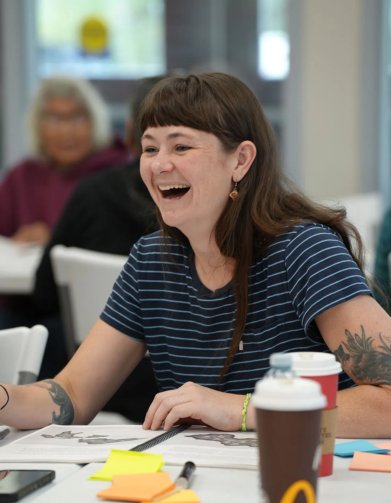 A woman with dark hair and bangs, wearing a blue and white striped t-shirt, laughs joyfully during a community meeting or workshop. She is seated at a table with an open spiral-bound notebook, colorful sticky notes, and coffee cups.