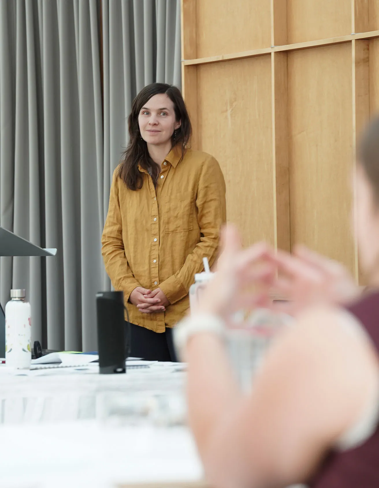 A woman with long dark hair, wearing a mustard-yellow button-down shirt, stands attentively at the front of a room during a training session. She is listening with a gentle, focused expression as a participant in the foreground gestures while speaking.