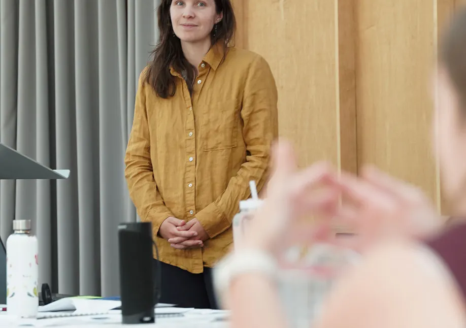 A woman with long dark hair, wearing a mustard-yellow button-down shirt, stands attentively at the front of a room during a training session. She is listening with a gentle, focused expression as a participant in the foreground gestures while speaking.