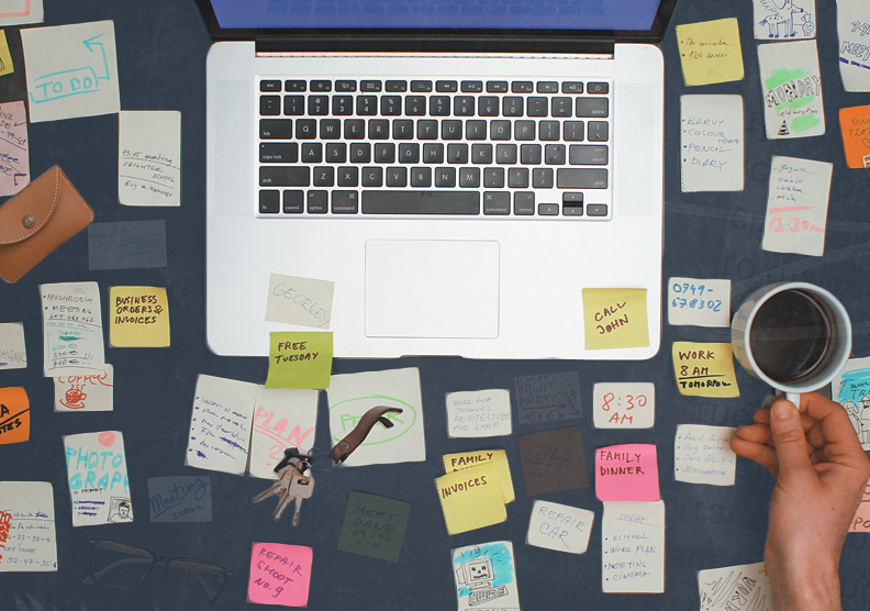 An overhead view of a desk with a computer open. The desk is filled with sticky notes and other objects showing reality of remote work.